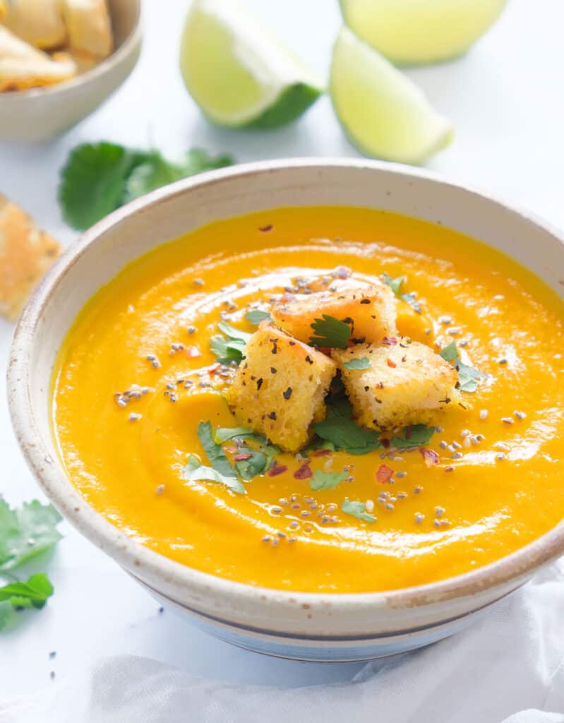 A bowl full of creamy carrot and ginger soup with croutons, slices of lime in the background.