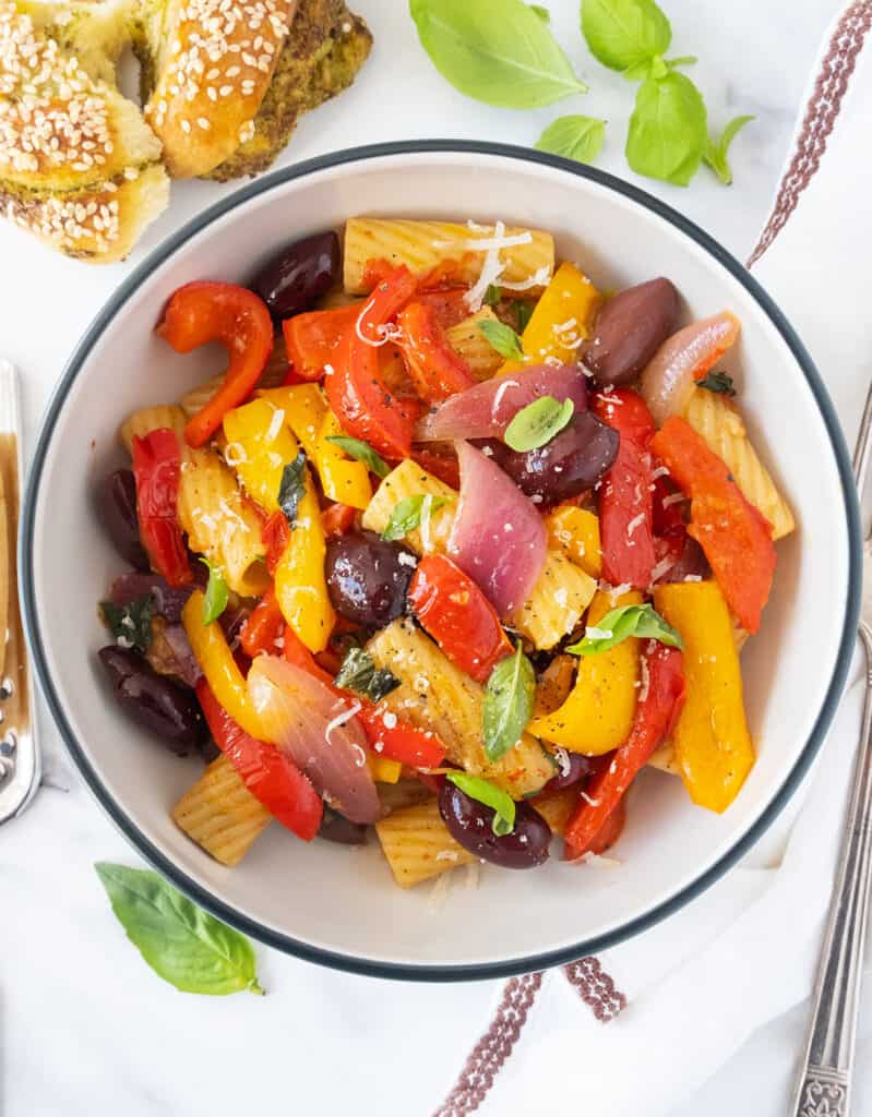 Top view of a white bowl full of pasta with peppers, black olives and basil leaves.