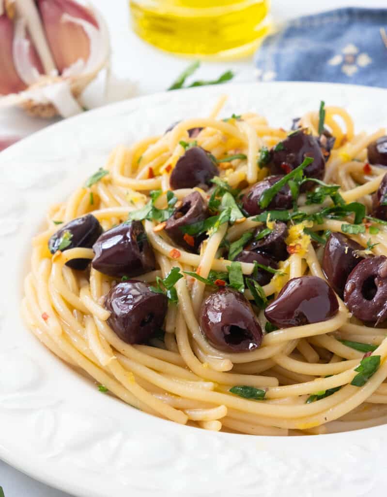 Close-up of a white plate full of pasta with olives and chopped parsley.