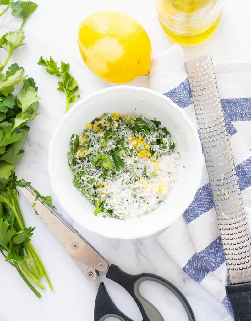 Top view of a small white bowl full of chopped parsley, lemon zest and grated parmesan cheese. A microplane, one lemon and a bunch of parsley in the background.