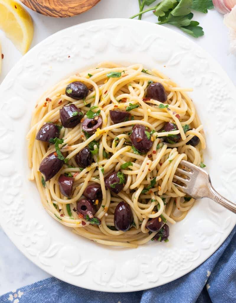 Top view of a white plate full of pasta with olives garnished with parsley and chilli flakes.