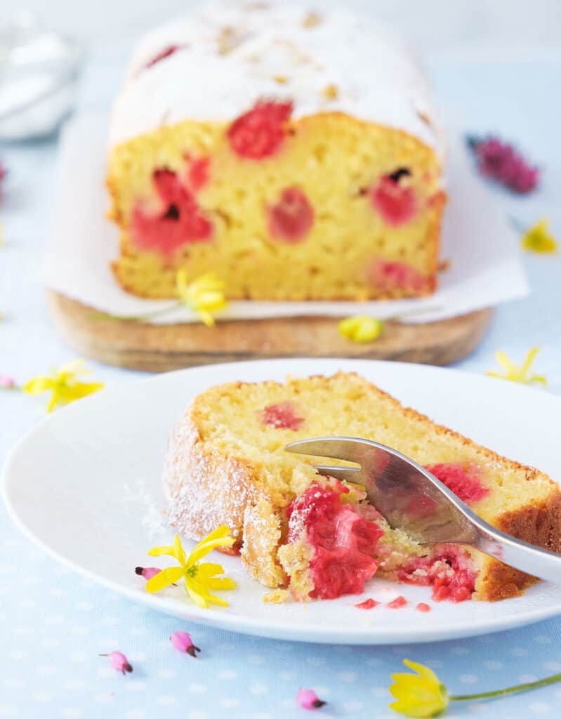 Close-up of a slice of raspberry ricotta cake with a for, small flowers and the whole cake loaf in the background.