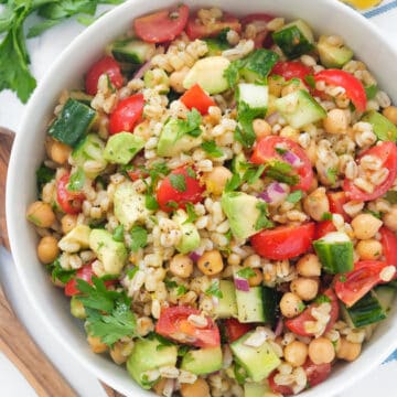 Top view of a white salad bowl full of barley salad with cherry tomatoes , diced cucumber and fresh herbs.
