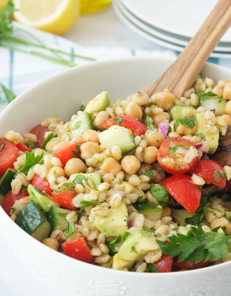 Close-up of a white salad bowl full of barley salad, lemons and white plates in the background.