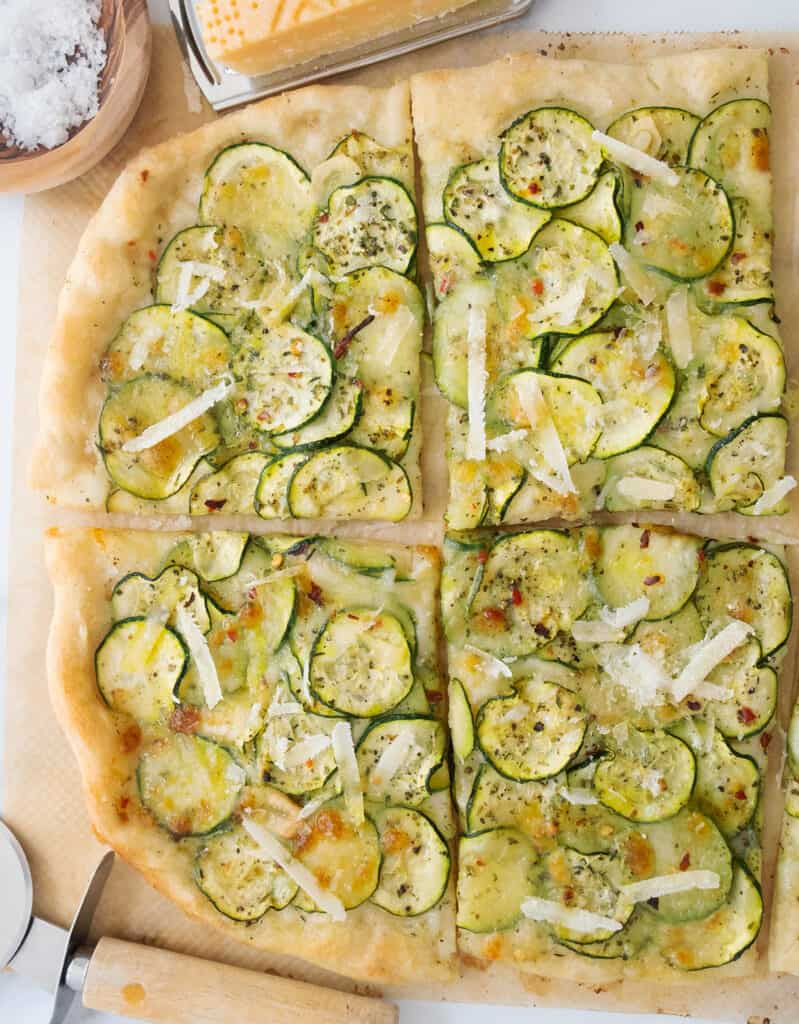 Top view of four slices of zucchini pizza garnished with shaved parmesan. Sea salt flakes, parmesan and a cheese greater in the background.