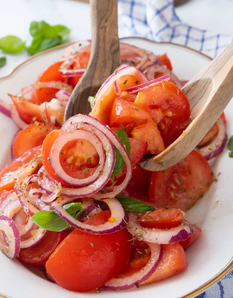 Two wooden spoons tossing tomato and onion salad in a white serving plate.