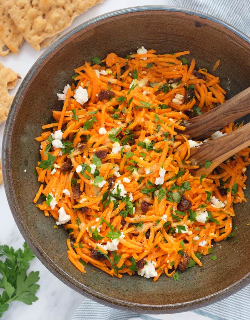 Top view of a brown bowl full of carrot raisin salad garnished with parsley and crumbled feta.