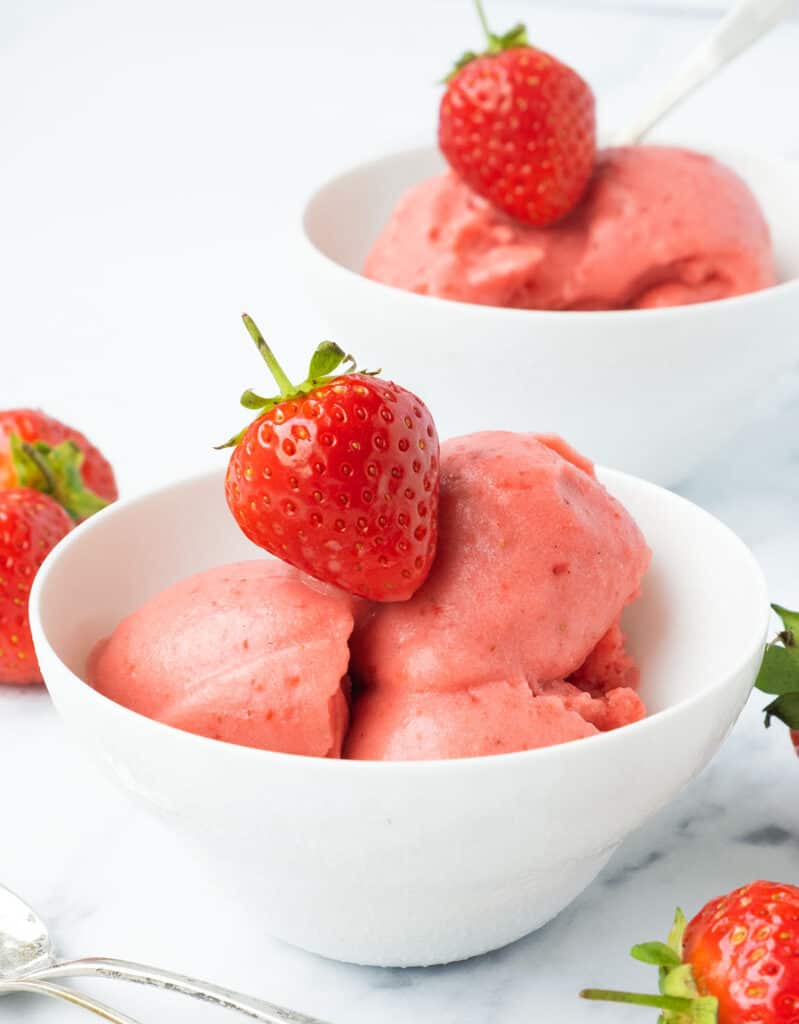 Two white bowls full of frozen strawberry yogurt topped with a fresh strawberry over a total white background.