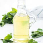 A small glass bottle full of mint syrup and mint leaves over a white background.