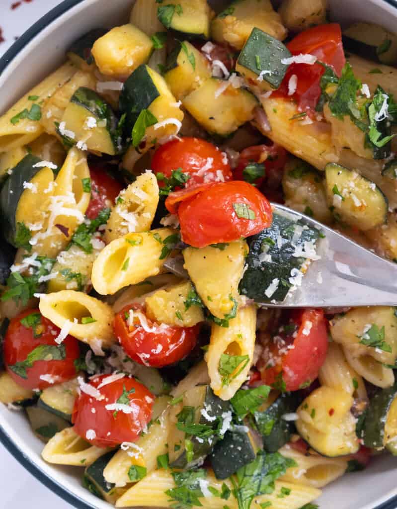 Close-up of a fork lifting some juicy pasta with tomatoes, zucchini and grated parmesan cheese.