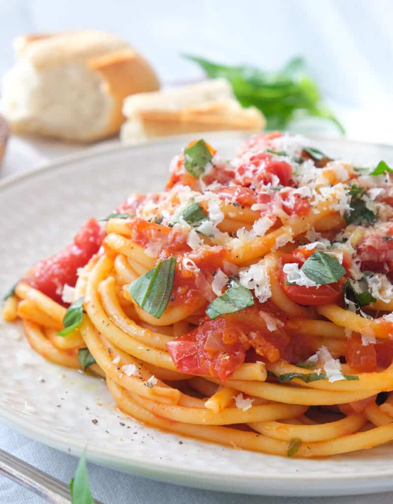 Close-up of a plate full of pasta napoletana with parmesan cheese and basil, fresh bread in the background.