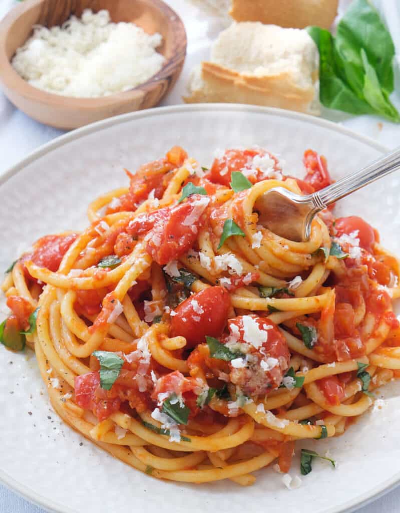 A white plate full of pasta alla napoletana, bread and grated parmesan cheese in the background.