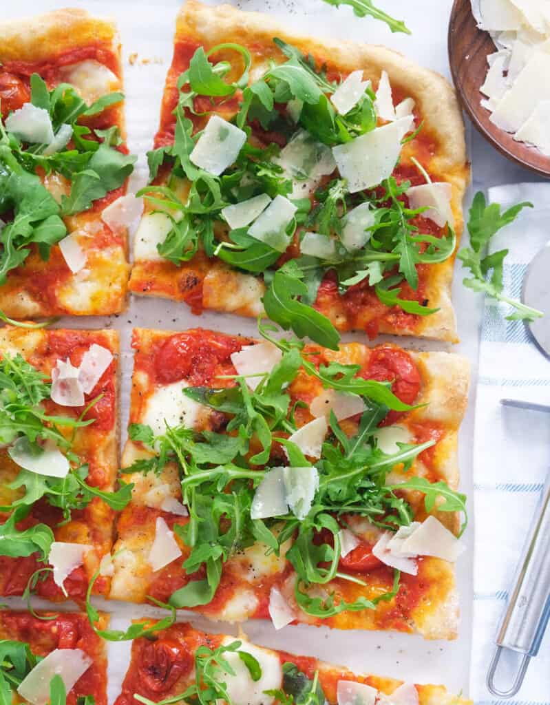 Top view of a pizza with arugula and shaved parmesan cut into slices over a white background.