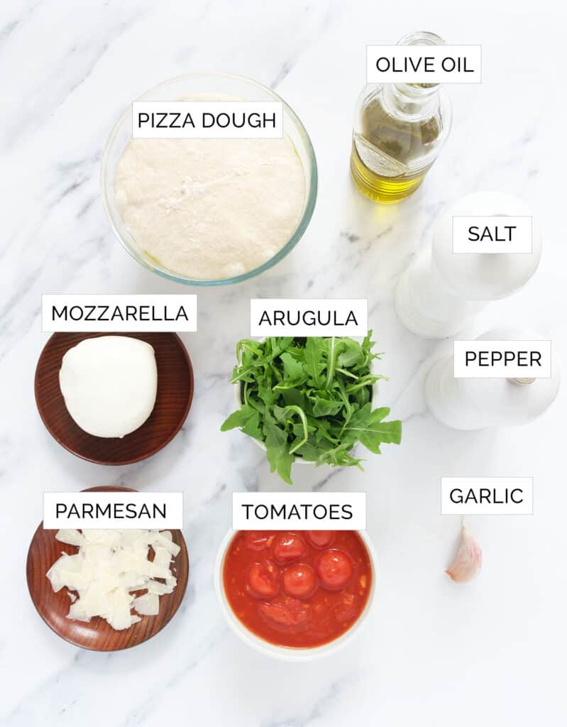 Top view of the ingredients to make this arugula pizza recipes are arranged over a white background.