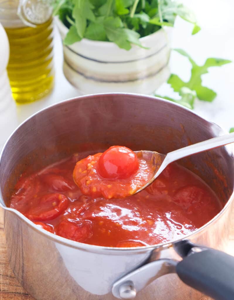 A spoon full of cherry tomato sauce, a sauce pan, a bottle with olive oil and fresh arugula in the background.