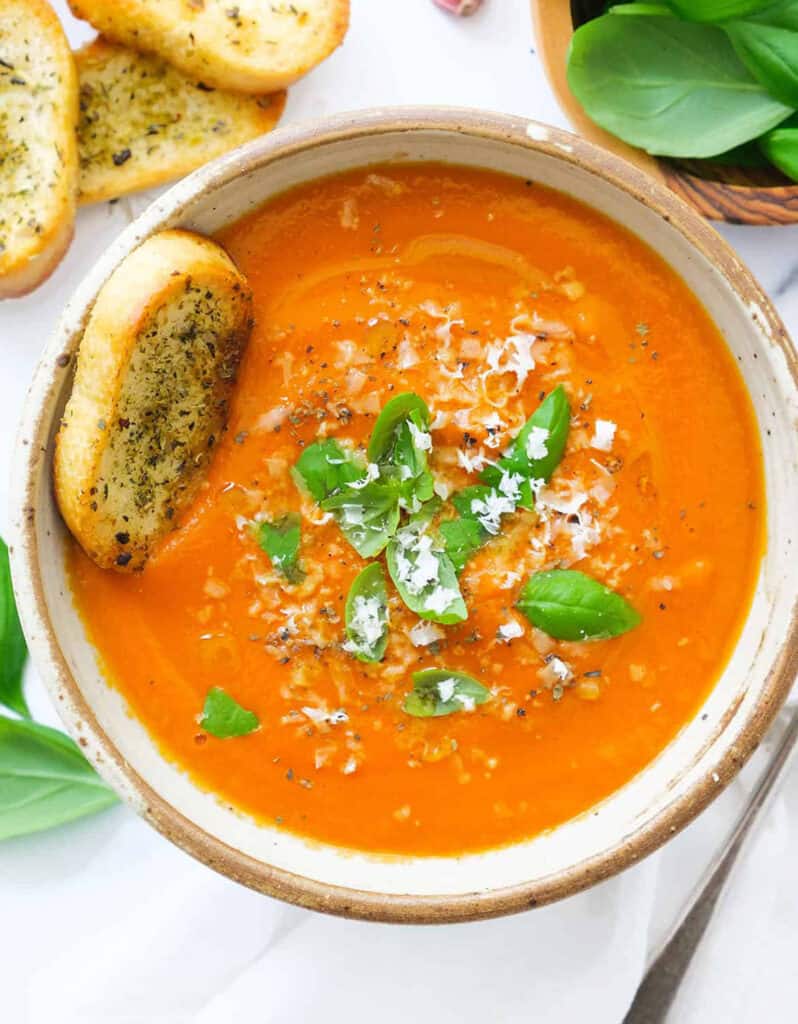 Top view of a bowl full of tomato soup with potatoes served with crusty bread and fresh basil.
