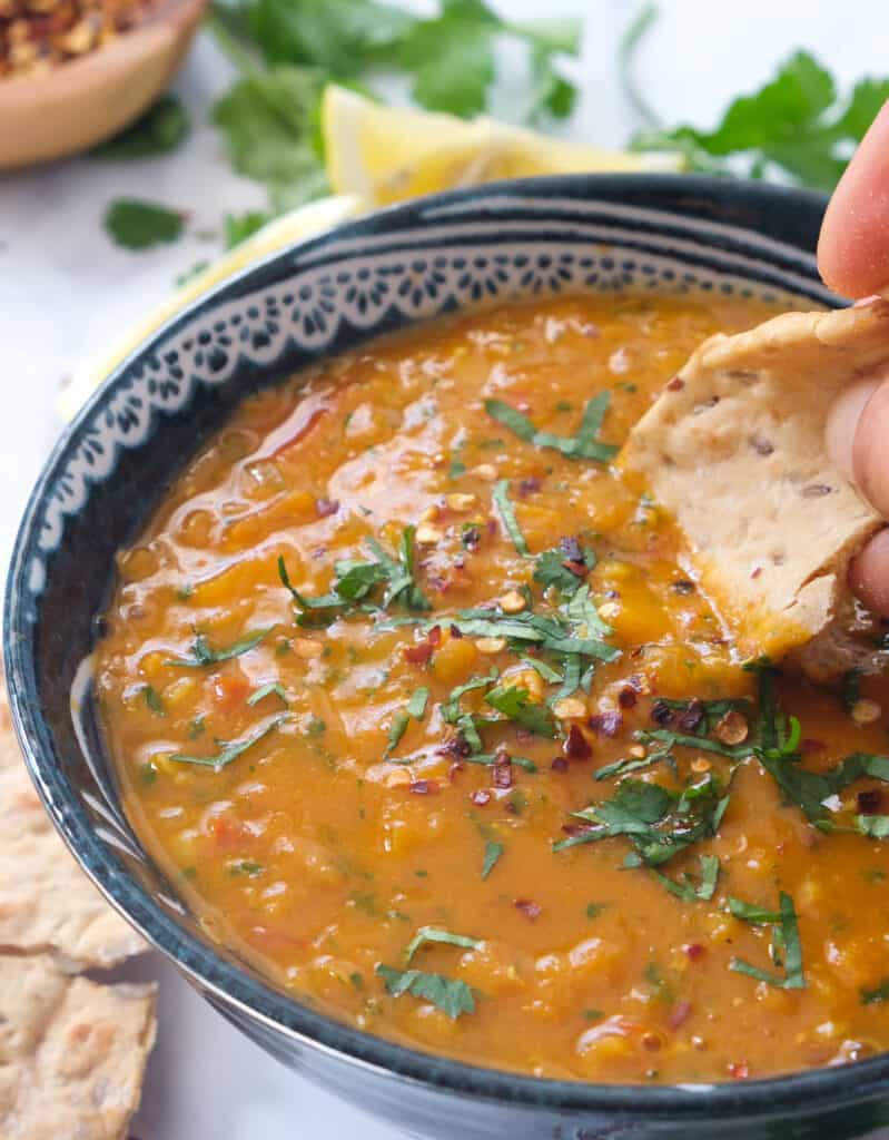 Close-up of a piece of pita bread dunking into a creamy tomato lentil soup garnished with fresh cilantro leaves.