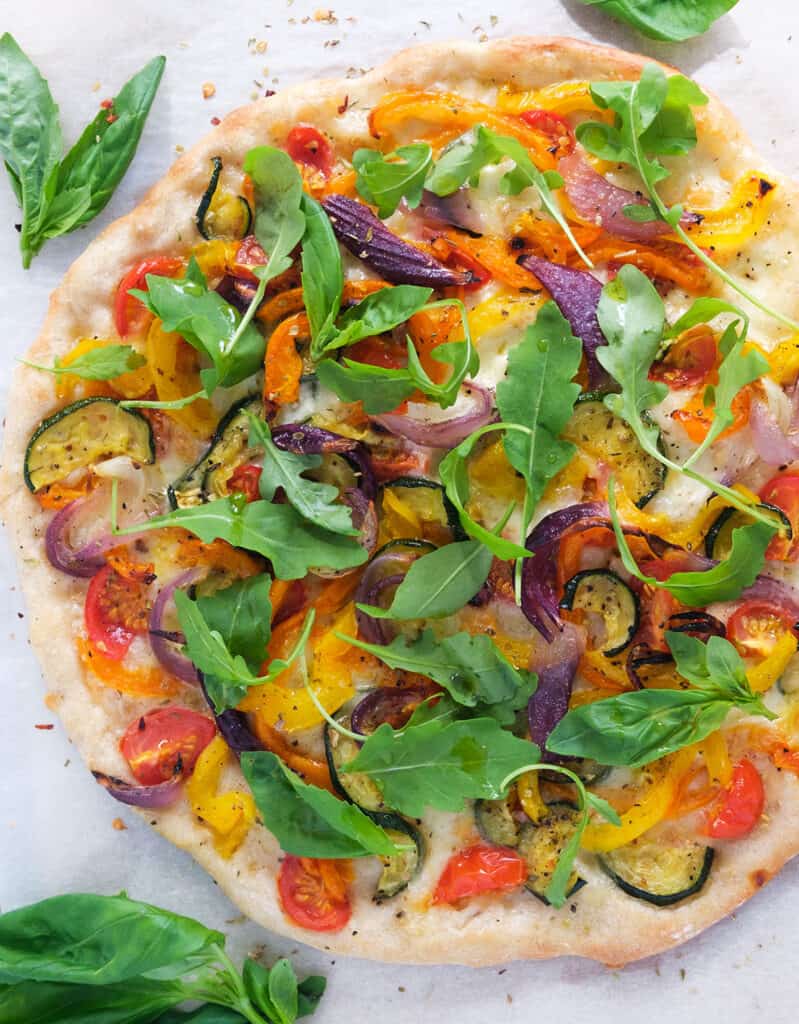 Top view of a veggie pizza full of roasted vegetables and arugula over a white background.