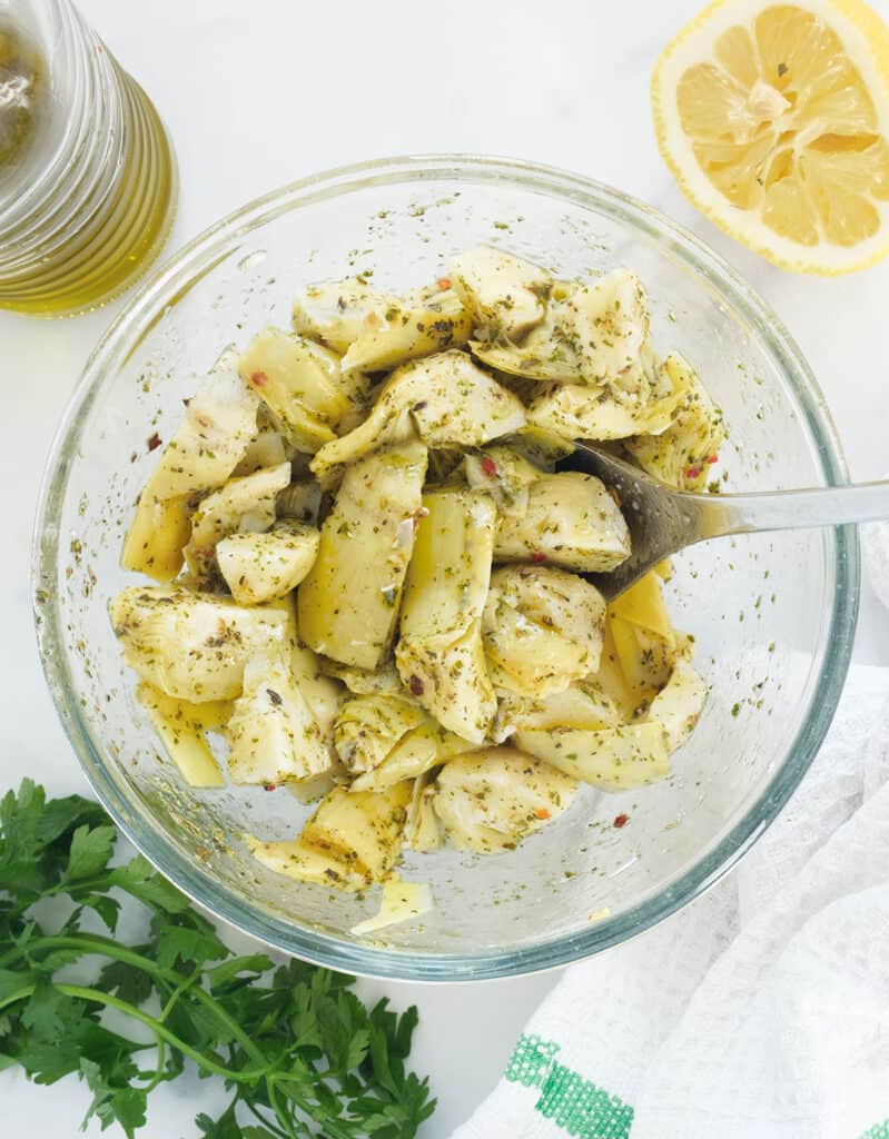 Top view of a glass bowl full of marinated artichokes, parsley and half lemon in the background.