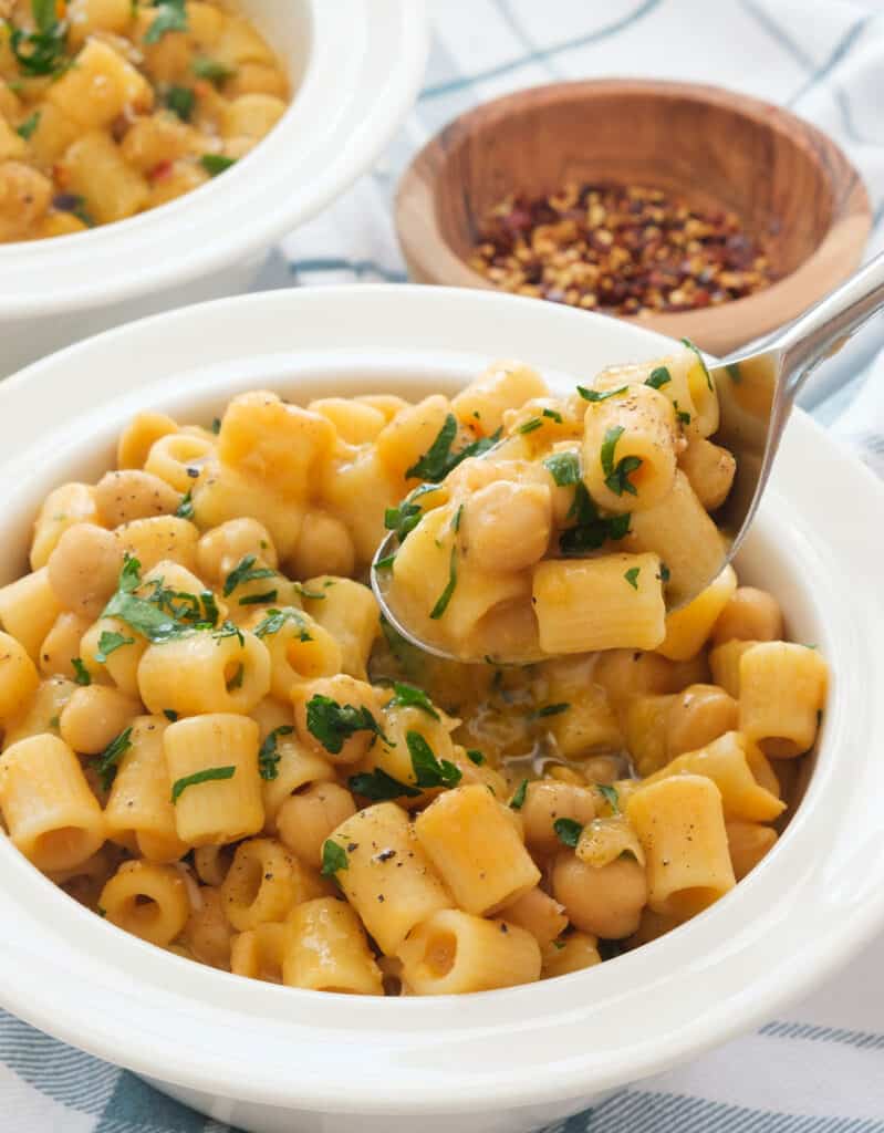 Close-up of a spoon full of pasta with chickpeas, a small wooden bowl full of chili flakes in the background.