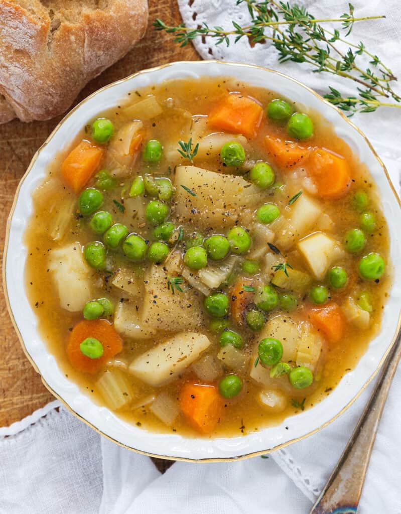 Top view of a white bowl full of potato stew with carrots and peas. Crusty bread and thyme in the background.
