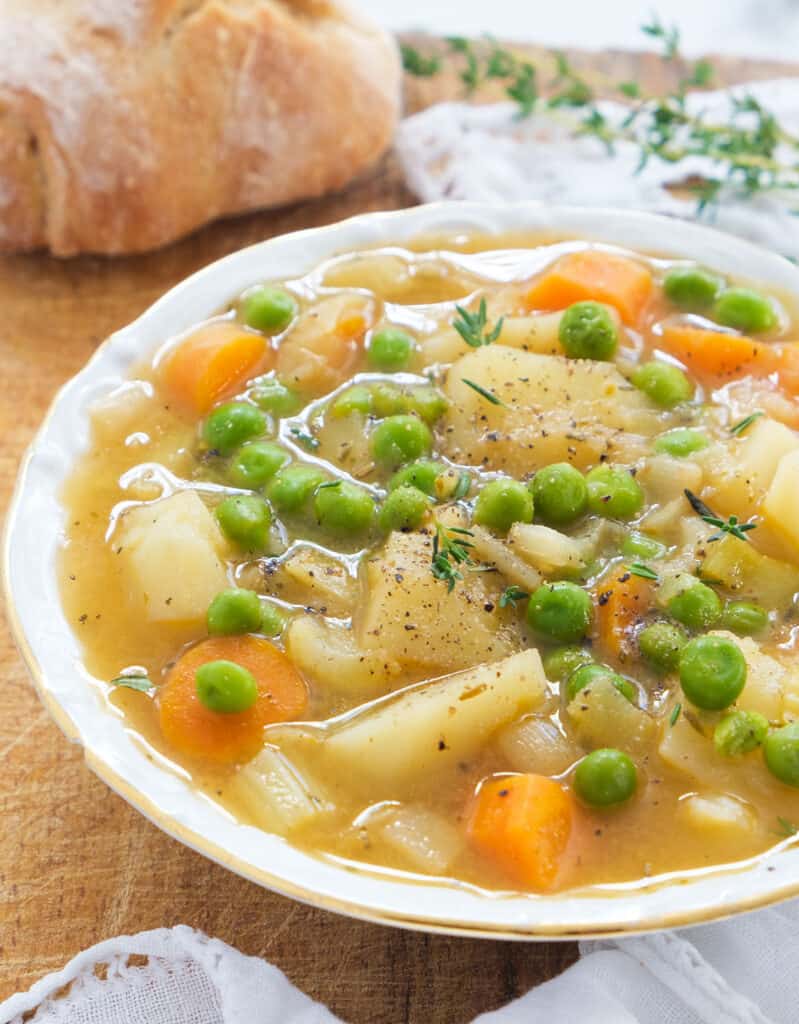 Close-up of a white bowl full of potato stew with peas, crusty bread in the background.