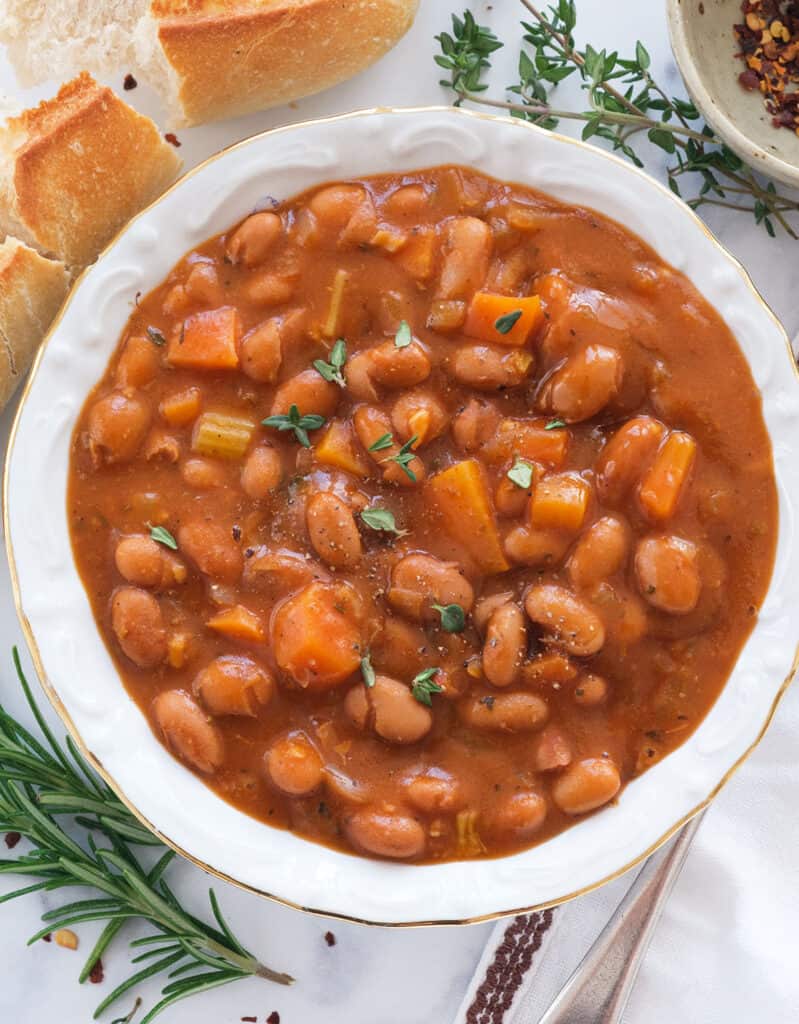 Top view of a white bowl full of bean stew, crusty bread and fresh thyme in the background.