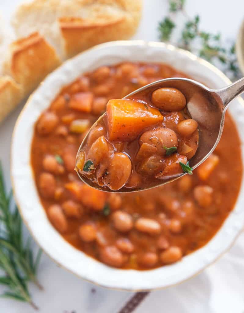 Close-up of a spoon full of beans cooked in a rich tomato sauce, a white bowl and some bread in the background.
