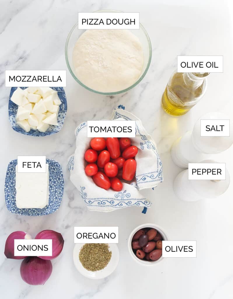 Top view of the ingredients to make this Greek pizza with feta cheese, a white marble surface in the background.