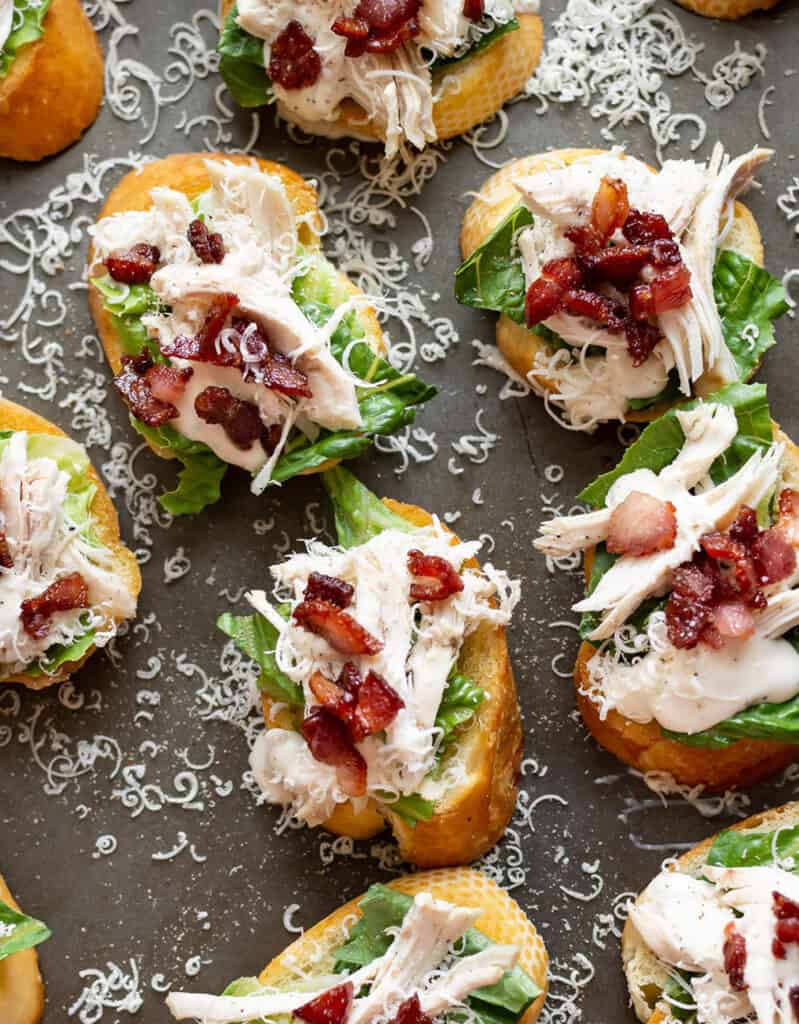 Top view of several crostini topped with chicken, crispy bacon, salad and grated parmesan.