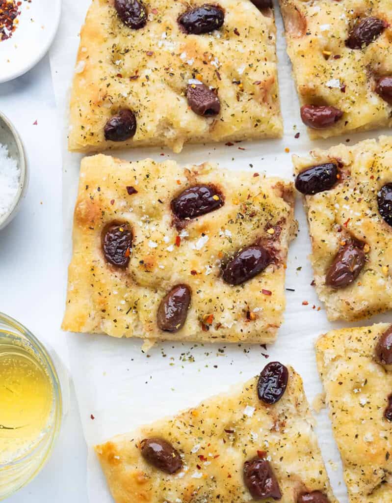 Top view of a large olive focaccia cut into squares and served over white parchment paper. Another great party food idea.