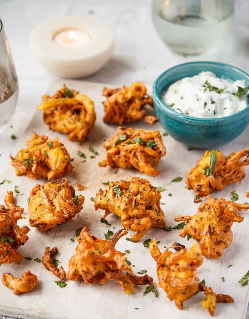 Several crispy onion fritters over a white back ground with a blue bowl full of minted yoghurt.