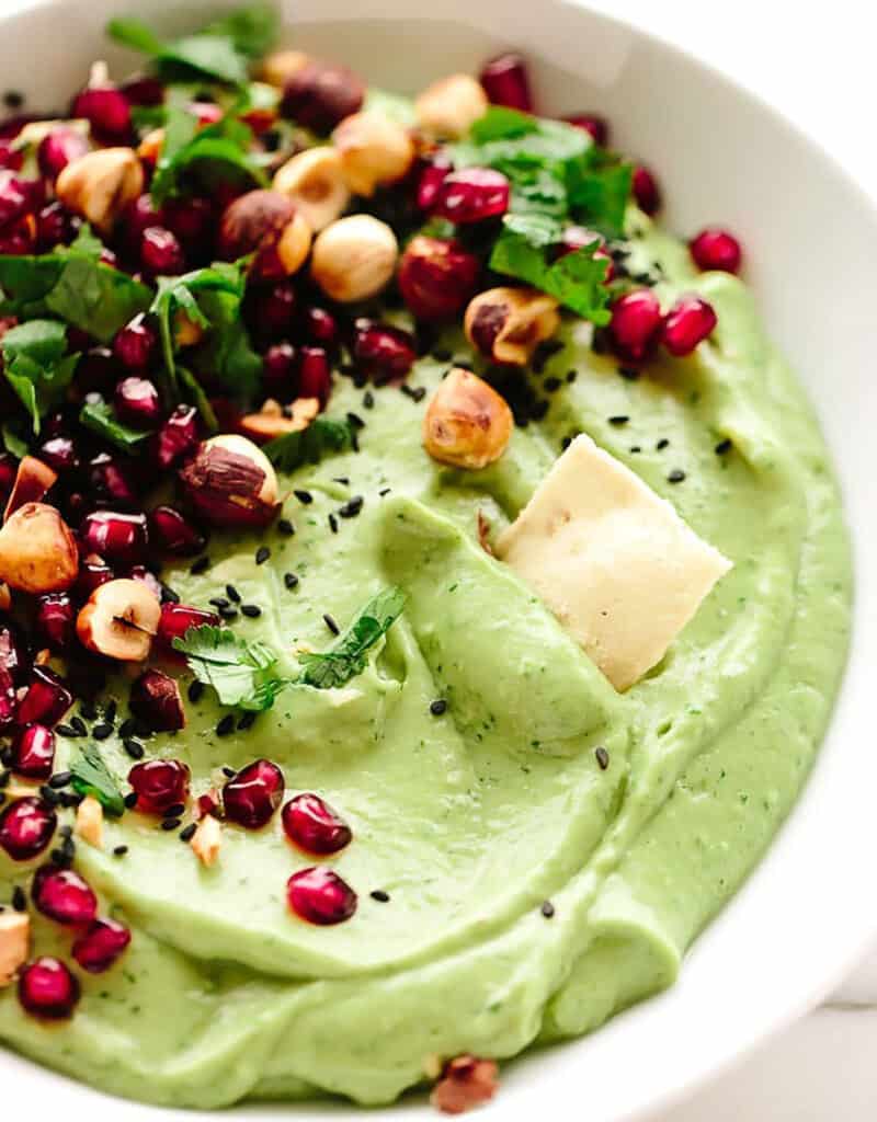 Close-up of a festive appetizer, a white bowl full of creamy avocado dip garnished with roasted hazelnuts and pomegranates.