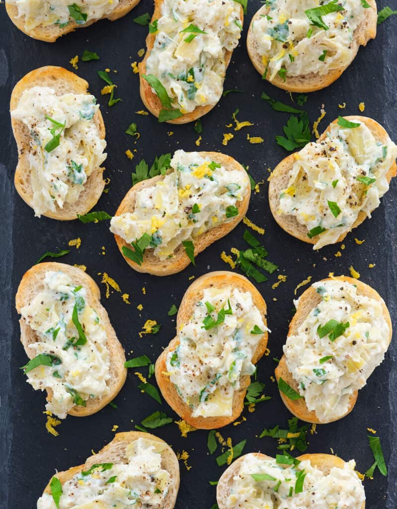 Top view of several servings of artichoke bruschetta garnished with parsley and lemon zest and served on a black tray.