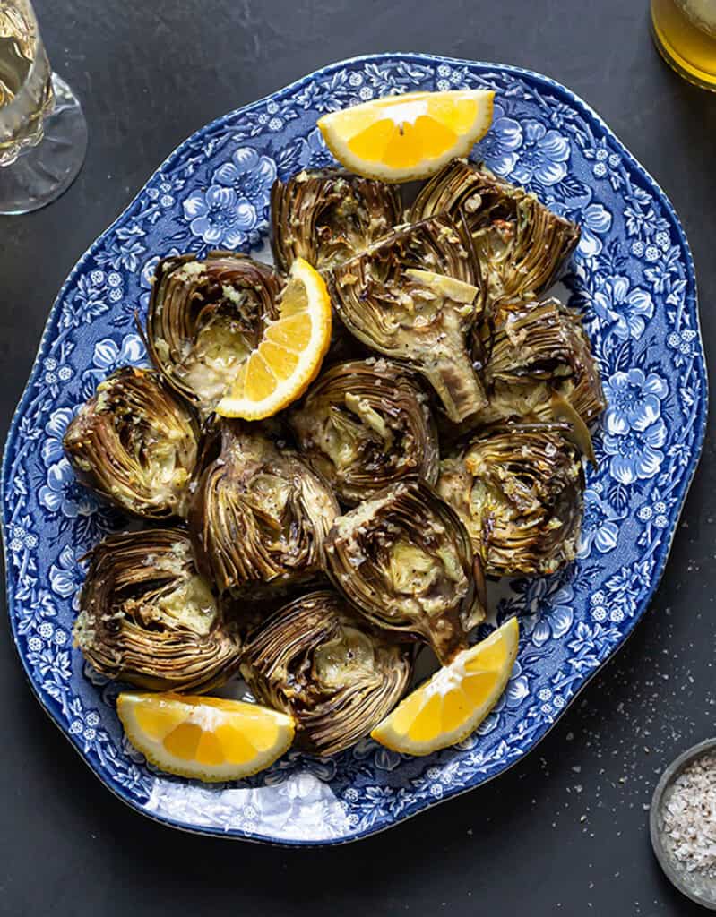 Top view of a blue tray full of roasted artichokes served with lemon wedges.