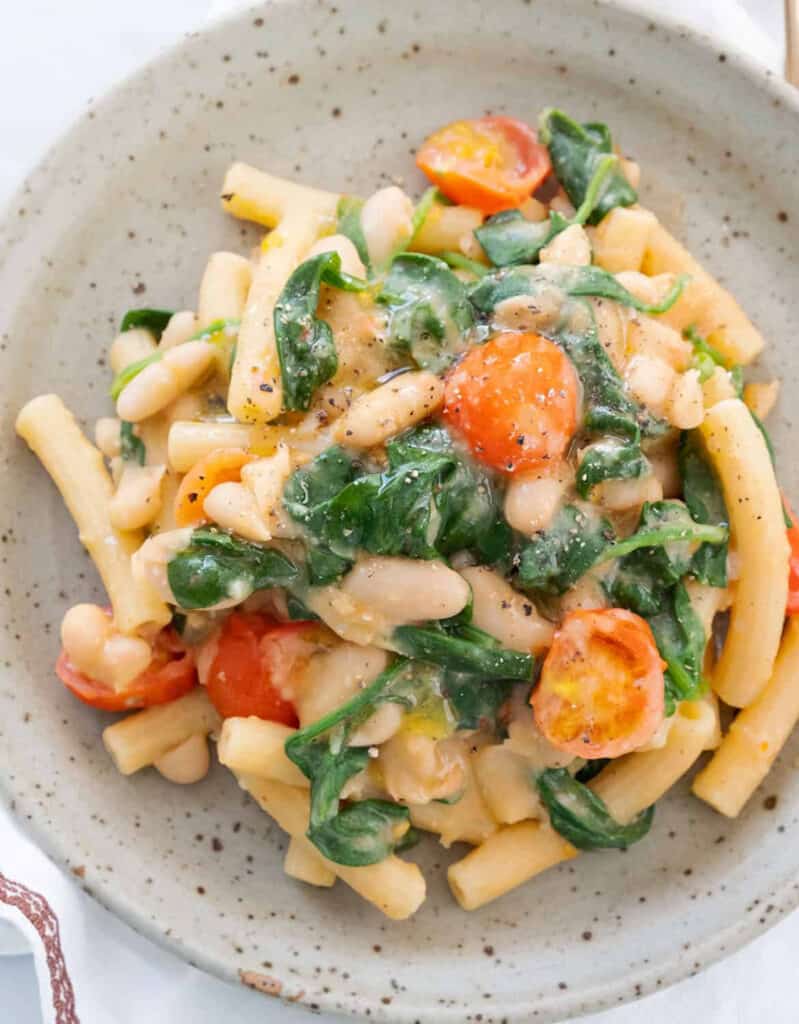 Top view of a grey plate with pasta with white beans, tomatoes and spinach.
