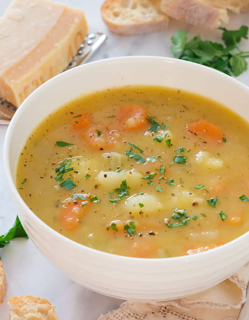 Close-up of a white bowl full of carrot potato soup served with ground pepper and chopped parsley.