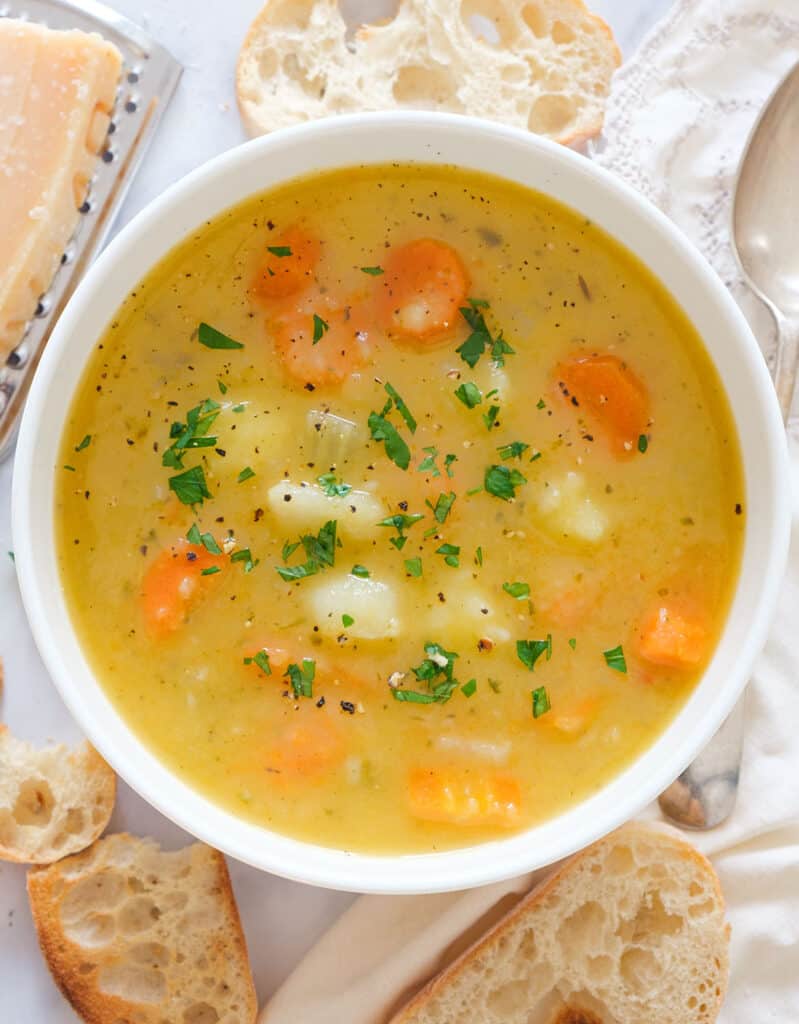 Top view of a white bowl full of carrot potato soup, slices of bread and parmesan cheese in the background.