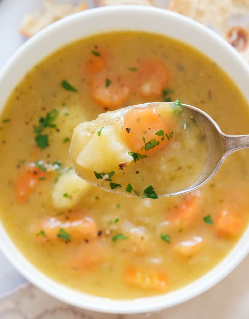 Close-up of a spoon full of carrot potato soup, and white bowl with soup in the background.