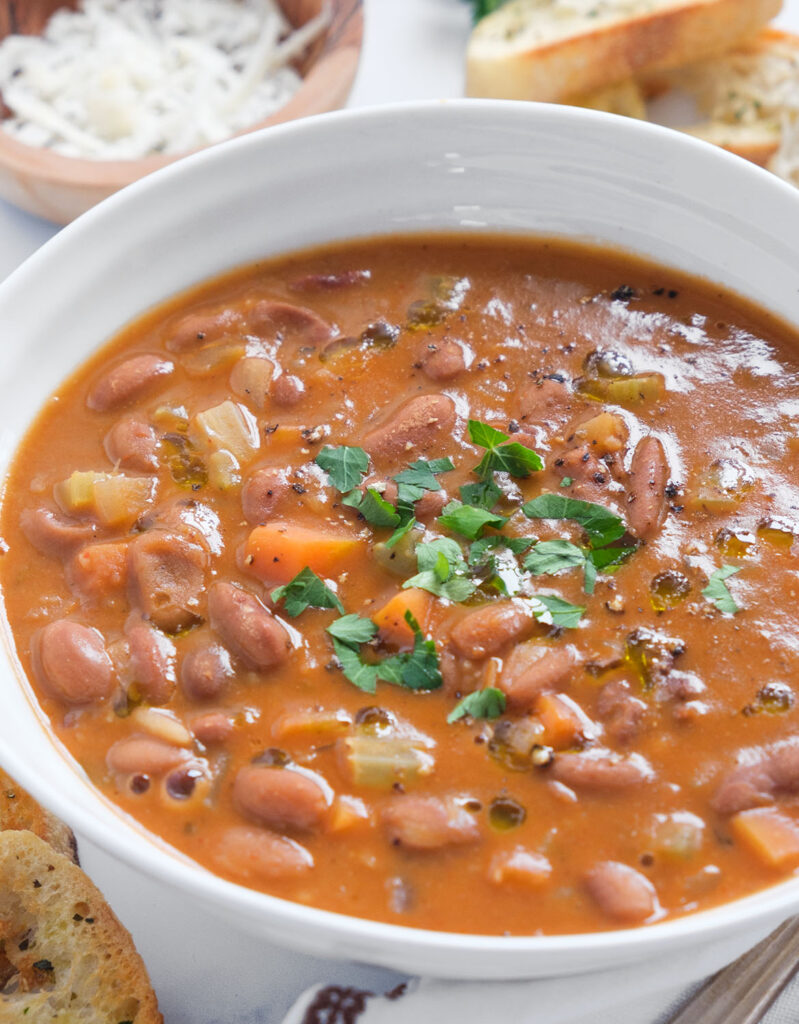 Close-up of a white bowl full of pinto bean soup, crusty bread in the background.