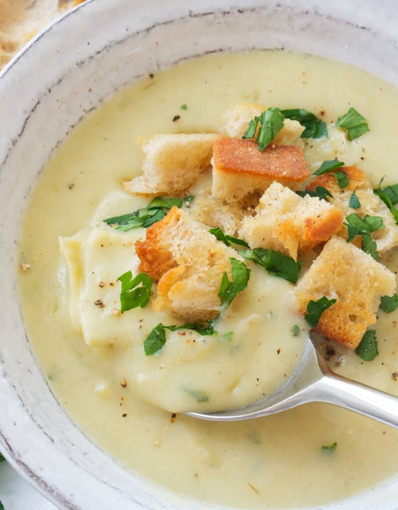 Close-up of a white bowl full of silky artichoke soup with crusty croutons and parsley.