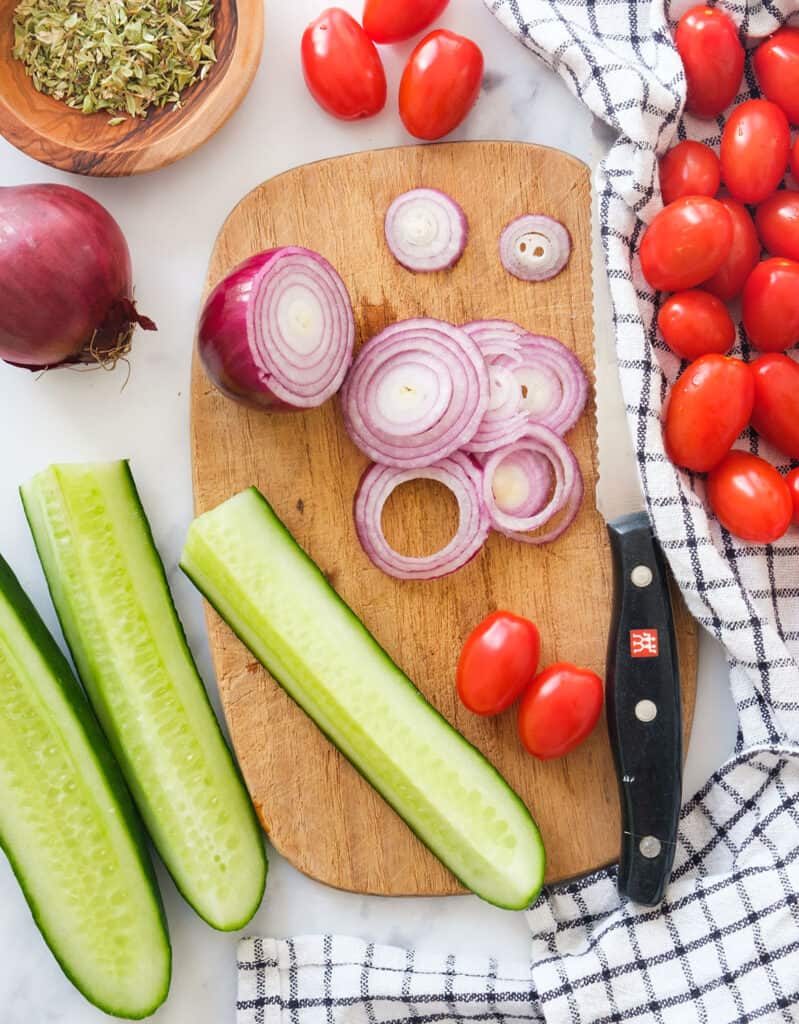Top view of a chopping board with sliced red onions, cucumber cut into quarters and cherry tomatoes.