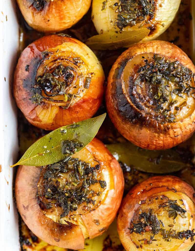 Top view of a baking pan full of roasted whole onions with bay leaves.