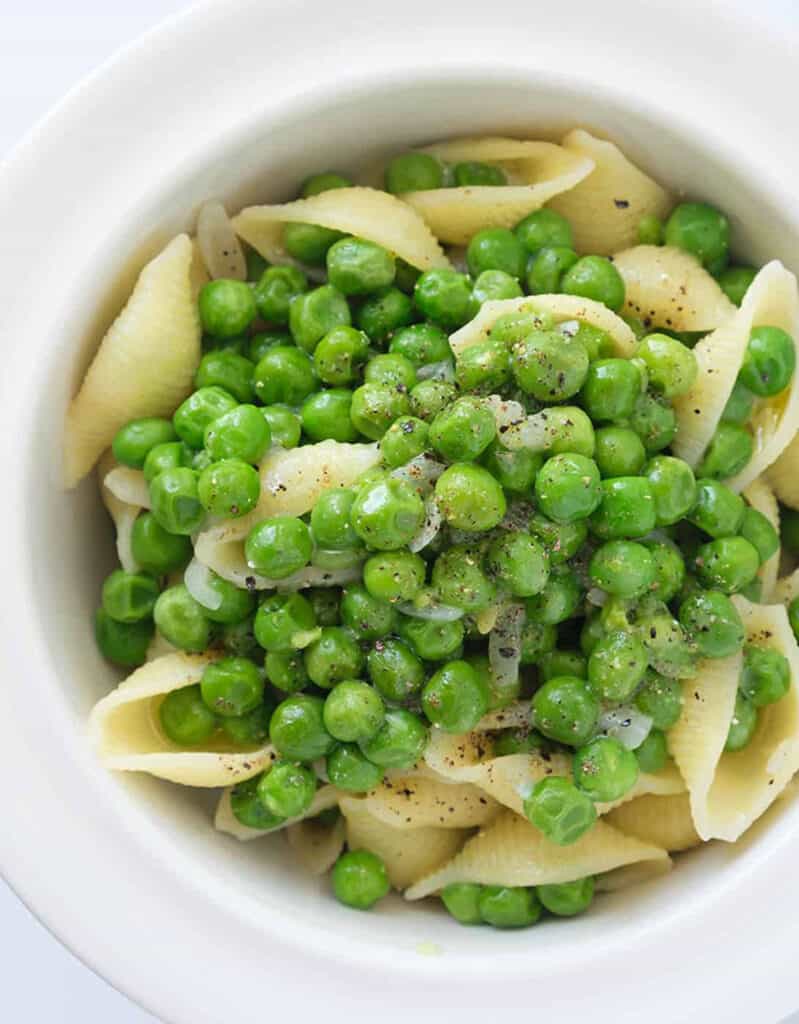 Top view of a white bowl full of pasta shells with green peas.