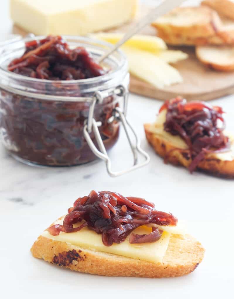 Crusty crostini with cheese and onion chutney and a glass jar full of chutney over a white background.