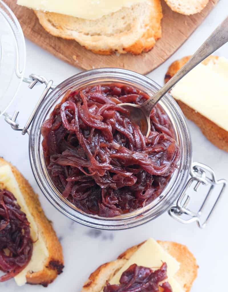 Top view of a glass jar full of red onion chutney and crostini with cheese over a white background.