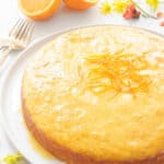 A round moist orange cake with oranges in the background.
