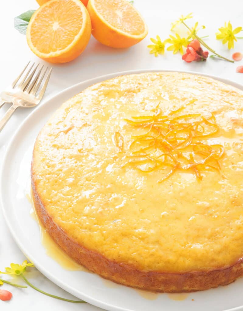 A round orange cake glazed with shiny syrup with oranges and small flowers in the background.
