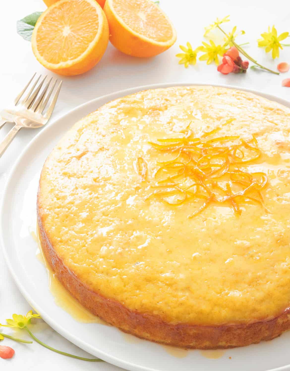 오렌지와 작은 꽃을 배경으로 반짝이는 시럽을 바른 둥근 오렌지 케이크.
