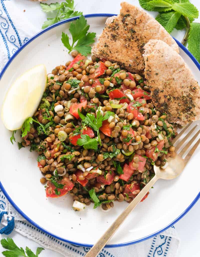 Top view of a white plate full of lentil tabbouleh served with a slice of lemon and pita bread.